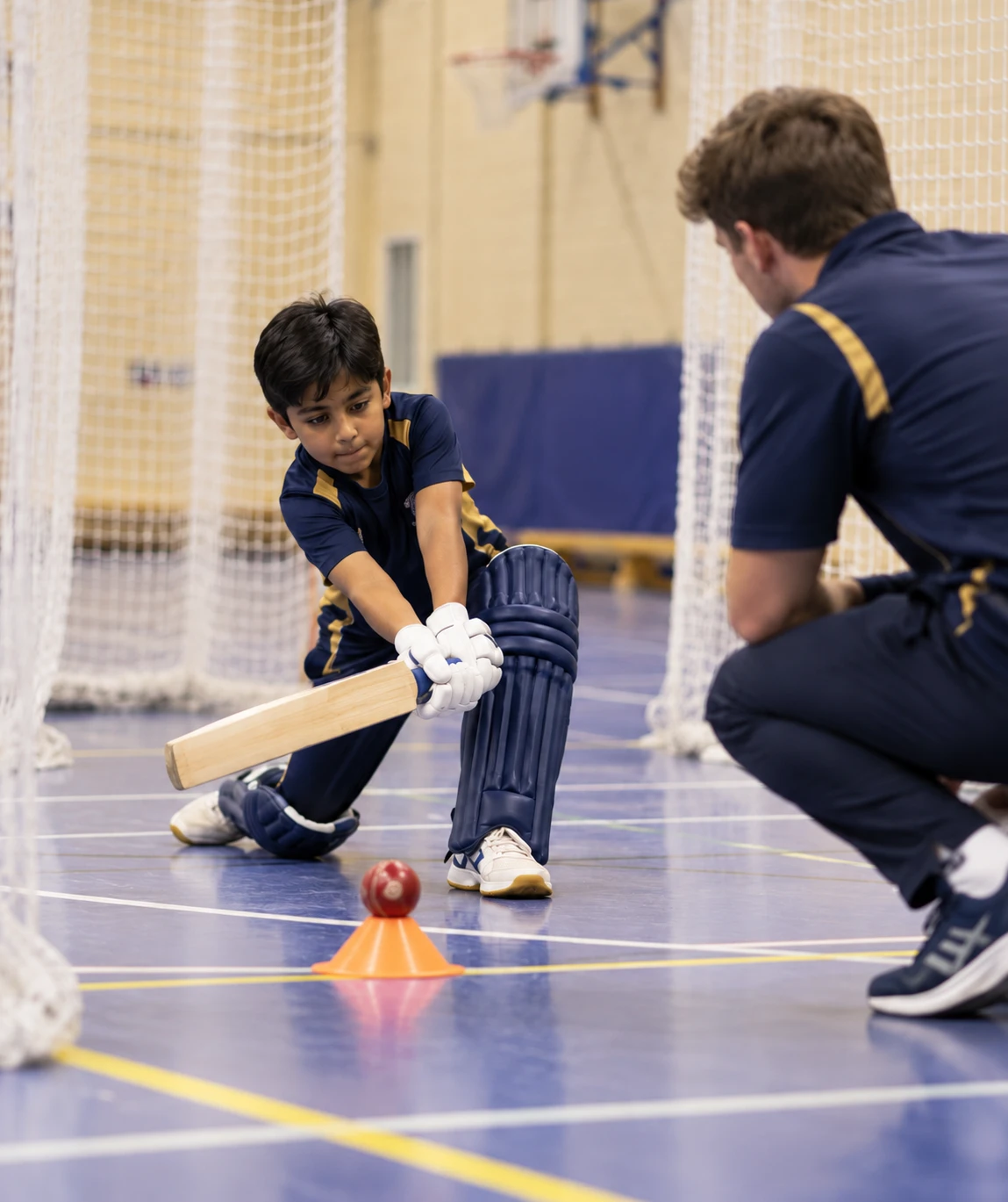 Coach watching a young batter practise a shot off a cone in an indoor net